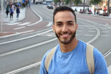 A smiling young Hispanic man with a beard wearing a blue t-shirt and backpack, showing a bright healthy smile in a city street setting for a DentaBiome customer testimonial.