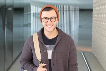 A smiling young man wearing glasses, an orange beanie, and a cardigan, showing a confident healthy smile for a DentaBiome customer product review.